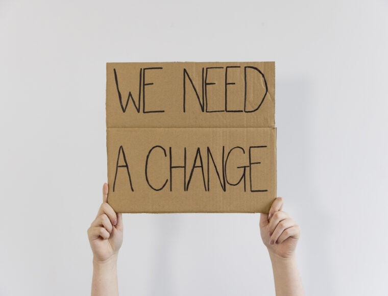 An unrecognisable person standing in front of a white background holding a handmade sign with 'WE NEED A CHANGE' on it up in the air.